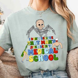 May include: A light green t-shirt with a colorful graphic. The graphic features a cartoon baby spider with the text "I DON'T BELIEVE THAT MAN'S EVER BEEN TO MEDICAL SCHOOL" in various colors.