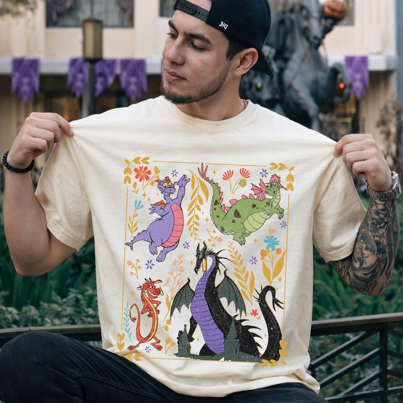 May include: Cream-colored t-shirt featuring a colorful dragon graphic. The design includes a purple dragon, a green dragon, a red dragon, and a black dragon, surrounded by floral elements within a gold border. The shirt is short-sleeved.