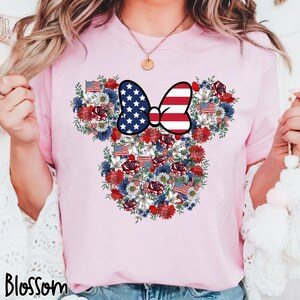May include: A light pink t-shirt featuring a floral design in the shape of a mouse head. The design incorporates red, white, and blue flowers, American flags, and a bow with a stars and stripes pattern. The word "Blossom" is printed at the bottom.