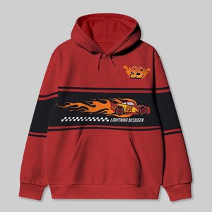 以下が含まれることがあります： 胸に黒と黄色のストライプが入った赤いフード付きスウェットシャツ。ストライプには、炎がついた赤いレーシングカーの漫画の画像と、「ライトニング・マックィーン」というテキストと「95」という数字が描かれています。