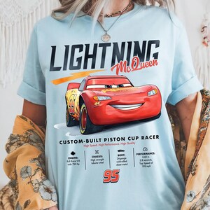 Puede incluir: Camiseta azul claro con un dise&ntilde;o de Rayo McQueen de la pel&iacute;cula Cars. El dise&ntilde;o incluye el texto "LIGHTNING McQueen" y "95", adem&aacute;s de las especificaciones del coche. La camiseta es un custom-built piston cup racer.