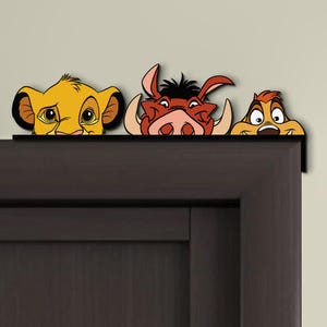 May include: A decorative door topper featuring Simba, Pumbaa, and Timon from The Lion King. The characters are in a cartoon style with vibrant colors, including yellow, brown, and red. The topper is designed to sit on top of a door frame.
