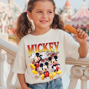 May include: A cream-coloured t-shirt with the text "MICKEY Mouse" in red and yellow lettering and three cartoon Mickey Mouse characters. The child is holding a churro.
