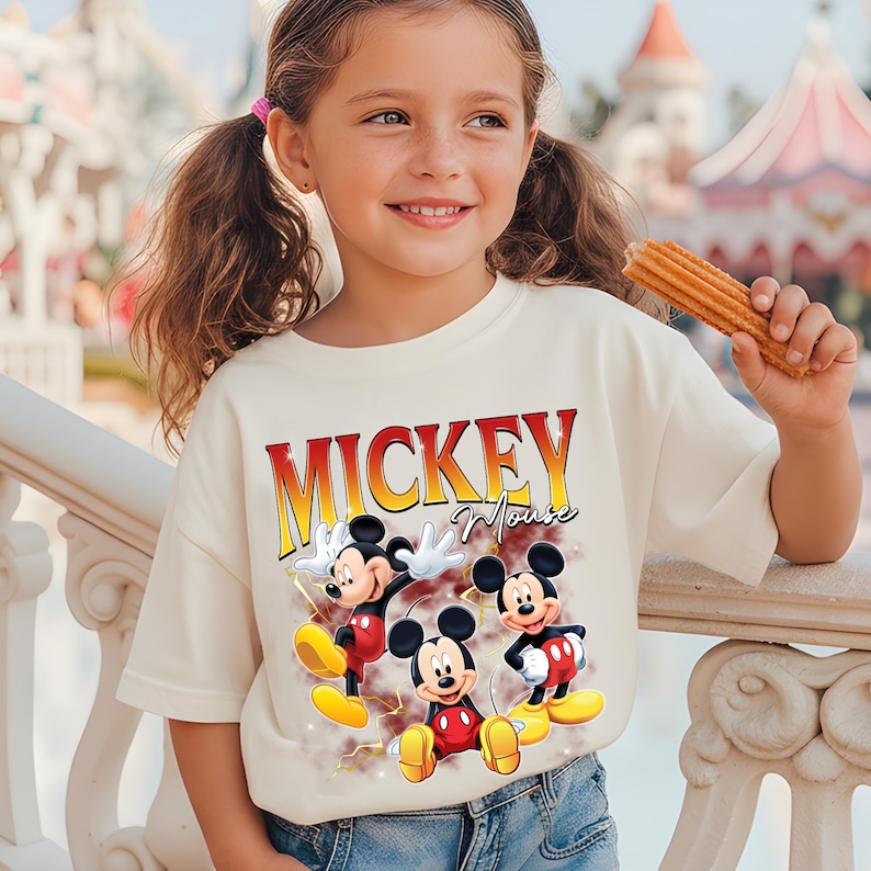 May include: A cream-coloured t-shirt with the text "MICKEY Mouse" in red and yellow lettering and three cartoon Mickey Mouse characters. The child is holding a churro.