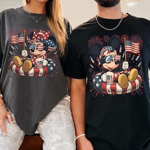 May include: Two t-shirts featuring Mickey and Minnie Mouse in patriotic designs. Each character is wearing sunglasses and holding an American flag and a mug. The shirts are dark gray and black, with a red, white, and blue color scheme.