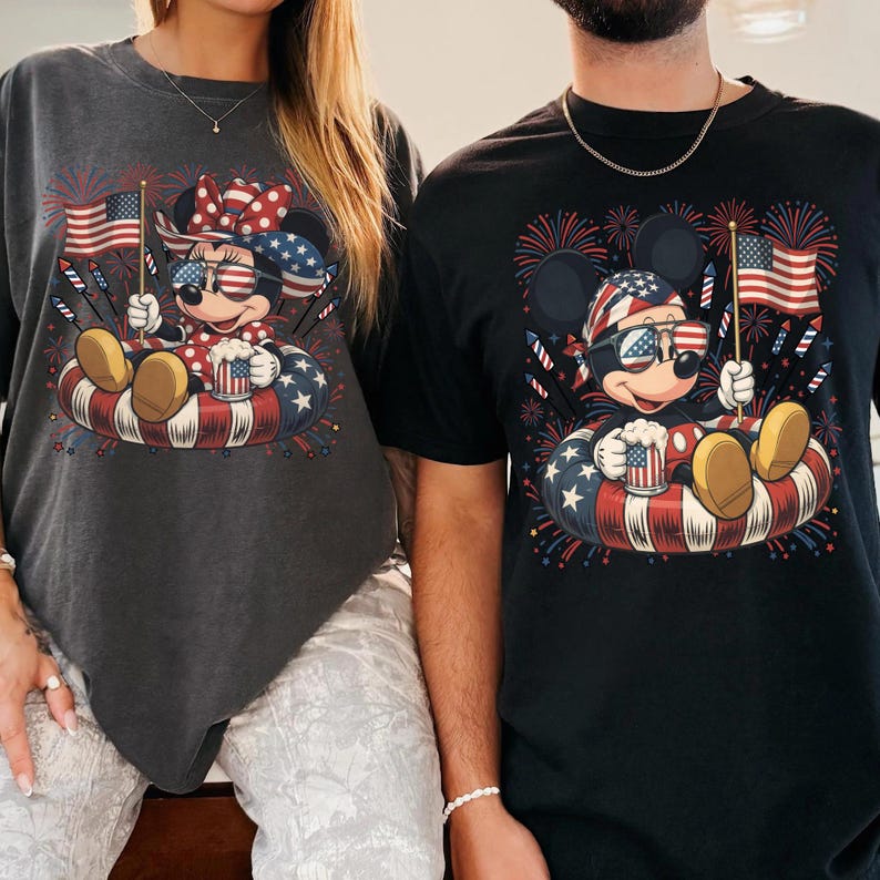 May include: Two t-shirts featuring Mickey and Minnie Mouse in patriotic designs. Each character is wearing sunglasses and holding an American flag and a mug. The shirts are dark gray and black, with a red, white, and blue color scheme.