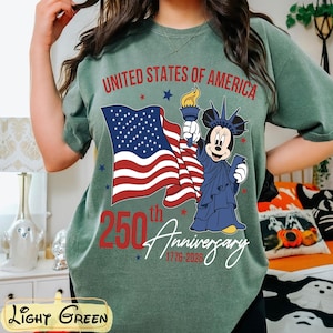 May include: Light green t-shirt with a graphic celebrating the 250th anniversary of the United States of America. The design includes the American flag, a cartoon character dressed as the Statue of Liberty, and the text "United States of America 250th Anniversary 1776-2026."