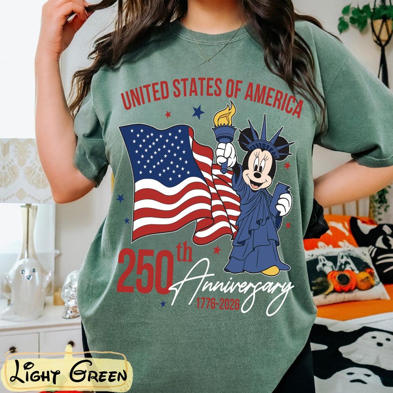 May include: Light green t-shirt with a graphic celebrating the 250th anniversary of the United States of America. The design includes the American flag, a cartoon character dressed as the Statue of Liberty, and the text "United States of America 250th Anniversary 1776-2026."