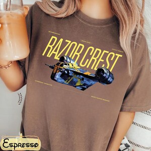 May include: Brown t-shirt featuring the text "RAZOR CREST" in yellow, with a detailed illustration of a spaceship. The shirt also includes the words "Decommissioned", "Mandalorian", "Assault Gunship", "Hyperspace Capabilities", and "Bounty Hunter Transport".