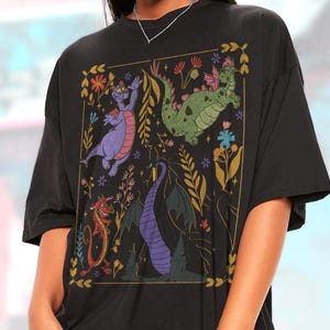 May include: Black t-shirt featuring a colorful graphic of cartoon dragons and floral elements within a gold rectangular frame. The dragons are in shades of purple, green, and red, with a variety of poses. The design includes flowers and foliage.