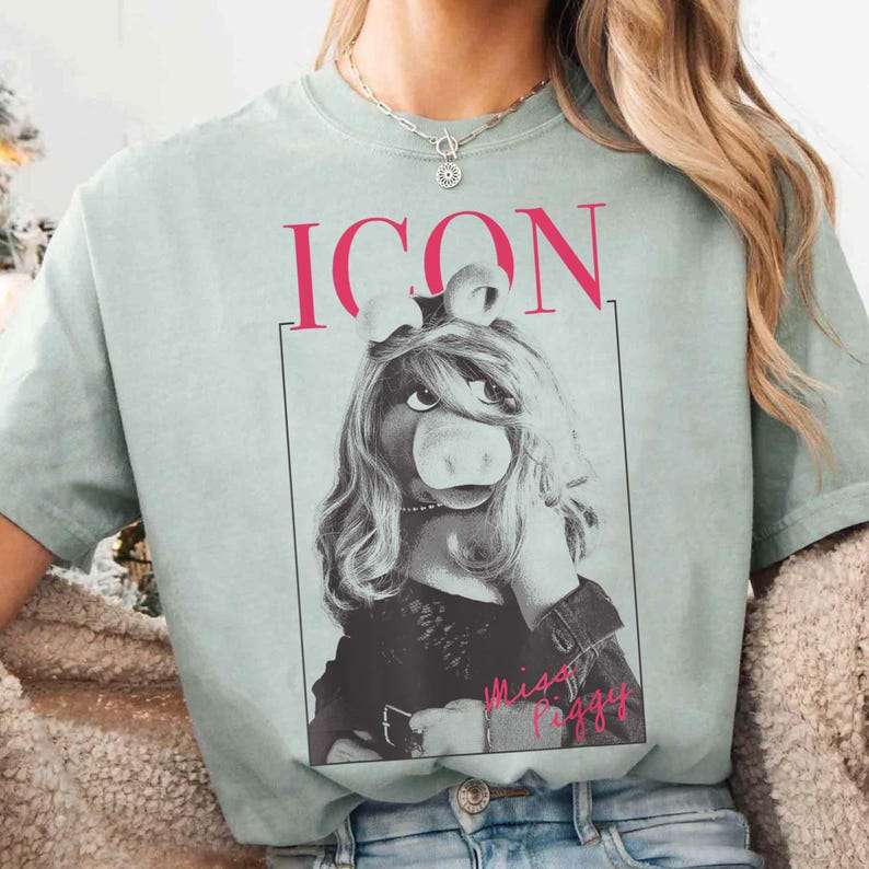 May include: A light green t-shirt featuring a black and white image of Miss Piggy from The Muppets. The word "ICON" is printed in pink above the image, and "Miss Piggy" is written in pink below.