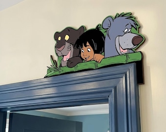 May include: A decorative wooden door topper featuring a colorful illustration of a black panther, a young boy with dark hair, and a grey bear peering over a green bamboo-style ledge. The cutout design is mounted on the top corner of a blue door frame.