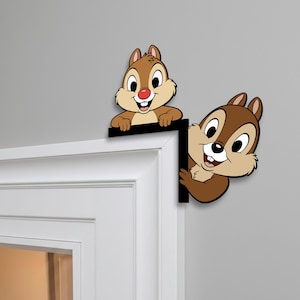 May include: Two cartoon squirrel figures, one brown and one tan, peek over a black corner piece. The squirrels have large eyes, red noses, and are smiling. The corner piece is mounted on a white door frame against a gray wall.