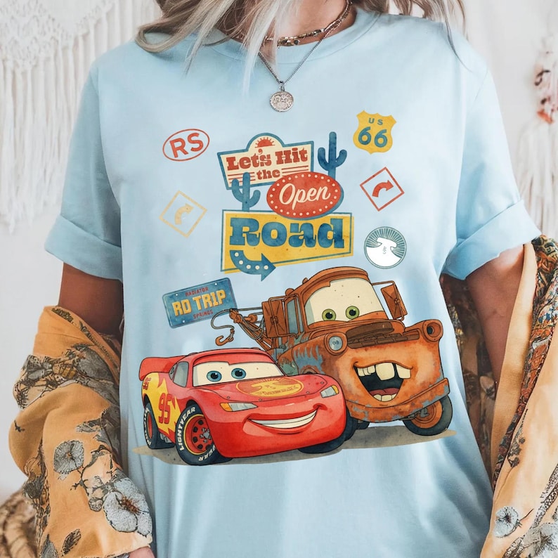 May include: Pale blue t-shirt with a graphic of Lightning McQueen and Mater from the film Cars. The design includes the text "Let's Hit the Open Road" and Route 66 signage. A bright, colourful design.