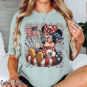 May include: Light blue t-shirt featuring a cartoon character wearing patriotic sunglasses and a bow, holding an American flag and a mug. The character is sitting in a red, white, and blue inner tube, with fireworks in the background. The text "Color Bay" is at the bottom.
