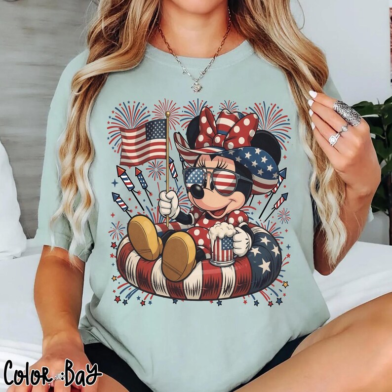 May include: Light blue t-shirt featuring a cartoon character wearing patriotic sunglasses and a bow, holding an American flag and a mug. The character is sitting in a red, white, and blue inner tube, with fireworks in the background. The text "Color Bay" is at the bottom.