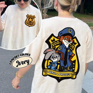 May include: Ivory t-shirt with a cartoon police badge on the front and a larger graphic on the back. The graphic shows a fox and a rabbit in police uniforms, with the text "Zootropolis Police" and "Serve & Protect".