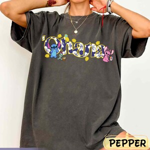 Puede incluir: Camiseta gris oscuro con la palabra "Ohana" en letras amarillas delineadas y un dise&ntilde;o floral. La camiseta presenta los personajes de dibujos animados Stitch y Angel, junto con elementos tropicales.