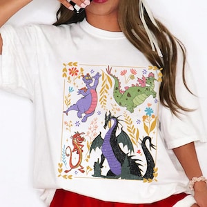 May include: White t-shirt featuring a colorful graphic of cartoon dragons. The design includes a purple dragon, a green dragon, a red dragon, and a black dragon, surrounded by flowers and leaves.