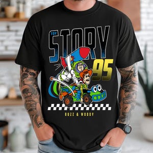May include: Black t-shirt featuring a colorful graphic of Buzz Lightyear and Woody from Toy Story in a race car. The design includes the text "TOY STORY 95" and "BUZZ & WOODY" in white and yellow. The shirt has a classic crew neck.