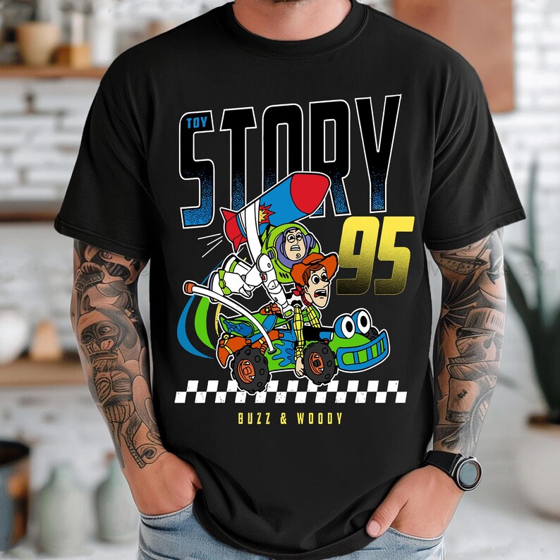 May include: Black t-shirt featuring a colorful graphic of Buzz Lightyear and Woody from Toy Story in a race car. The design includes the text "TOY STORY 95" and "BUZZ & WOODY" in white and yellow. The shirt has a classic crew neck.