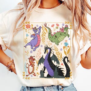May include: Cream-colored sweatshirt featuring a colorful dragon graphic. The design includes a purple dragon, a green dragon, a red dragon, and a black dragon, surrounded by floral elements. The graphic is framed within a rectangular border.