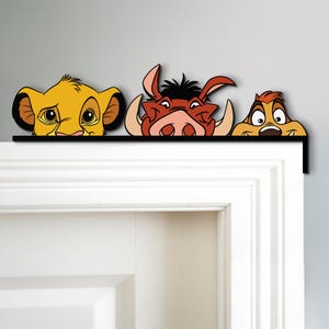 May include: A decorative door topper featuring Simba, Pumbaa, and Timon from The Lion King. The characters are in vibrant colors, with Simba in yellow, Pumbaa in reddish-brown, and Timon in tan. The topper is mounted on a black bar.