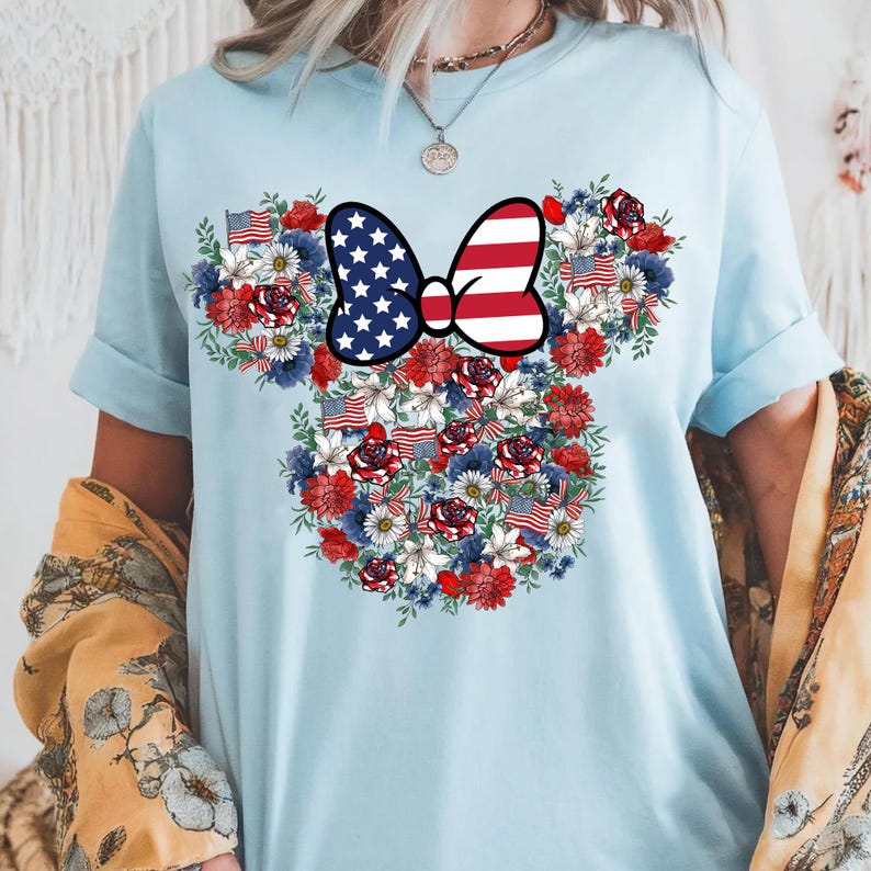 May include: Light blue t-shirt featuring a floral design in the shape of a mouse head. The design includes red roses, white daisies, blue flowers, and American flags. A bow with a stars and stripes pattern is at the center.