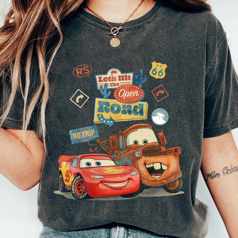May include: Dark grey t-shirt with a graphic of Lightning McQueen and Mater from the film Cars, and the text "Let's Hit the Open Road". The design includes road trip signs. The shirt has a vintage, worn appearance.