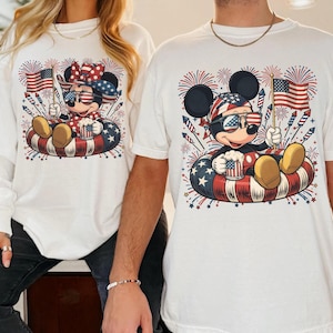 May include: White t-shirts featuring Mickey and Minnie Mouse in patriotic designs. Mickey and Minnie are wearing sunglasses and holding American flags, sitting in an inner tube with red, white, and blue stars and stripes. Fireworks are in the background.