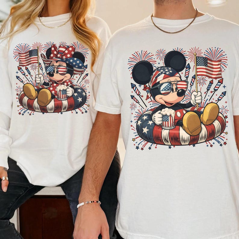 May include: White t-shirts featuring Mickey and Minnie Mouse in patriotic designs. Mickey and Minnie are wearing sunglasses and holding American flags, sitting in an inner tube with red, white, and blue stars and stripes. Fireworks are in the background.