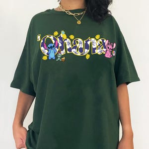 Puede incluir: Camiseta verde oscuro con la palabra "Ohana" en un dise&ntilde;o floral y personajes de dibujos animados. La camiseta tiene un corte holgado y mangas cortas. Los personajes incluyen un extraterrestre azul y un extraterrestre rosa.