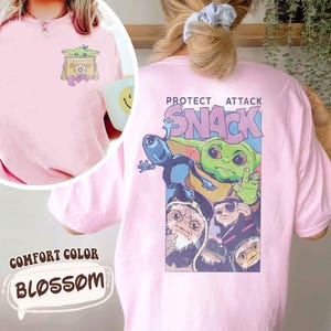 May include: A light pink comfort color t-shirt with a graphic on the front and back. The front graphic features a cartoon character in a box with flowers. The back graphic says "PROTECT ATTACK SNACK" with cartoon characters.