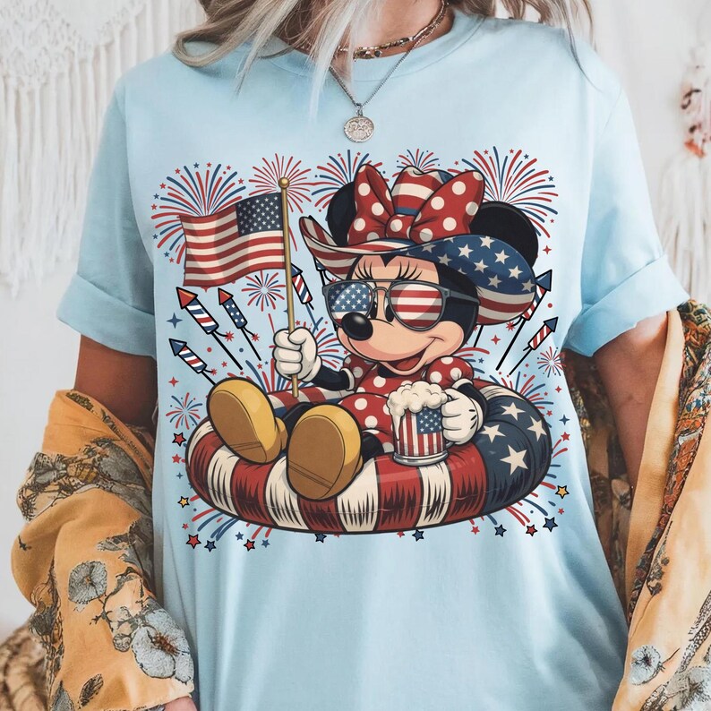 May include: Light blue t-shirt featuring a cartoon mouse wearing sunglasses, a patriotic bow, and holding a mug and American flag. The mouse is sitting on a red, white, and blue inner tube, with fireworks in the background.