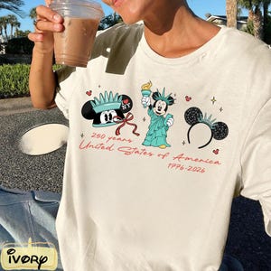 May include: Off-white t-shirt featuring a graphic of Mickey Mouse in a Statue of Liberty costume, a Mickey Mouse hat, and a Mickey Mouse ear headband. The text "250 years United States of America 1776-2026" is printed in red.