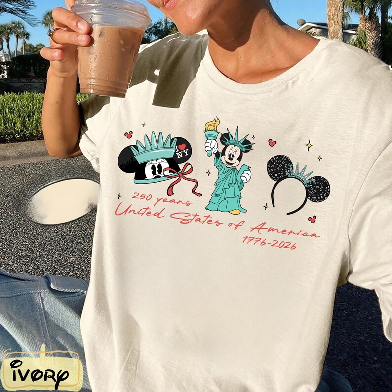 May include: Off-white t-shirt featuring a graphic of Mickey Mouse in a Statue of Liberty costume, a Mickey Mouse hat, and a Mickey Mouse ear headband. The text "250 years United States of America 1776-2026" is printed in red.