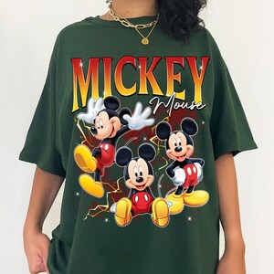 May include: Dark green t-shirt with a Mickey Mouse graphic. The design includes the word "MICKEY" in red and yellow, above "Mouse" in script. Mickey is depicted in several poses, wearing red shorts and yellow shoes.