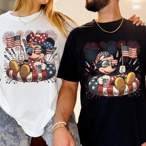 May include: White and black t-shirts featuring a cartoon mouse character in patriotic attire. The mouse is wearing sunglasses and holding an American flag and a mug. The design includes fireworks and a red, white, and blue inner tube.