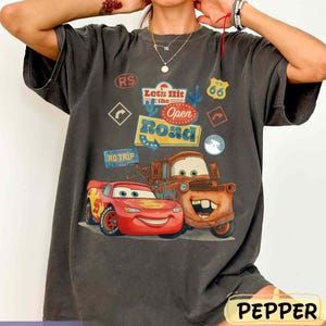 May include: Dark grey t-shirt featuring Lightning McQueen and Mater from the film Cars, with the text "Let's Hit the Open Road" and Route 66 signage. The shirt has a vintage, worn appearance.