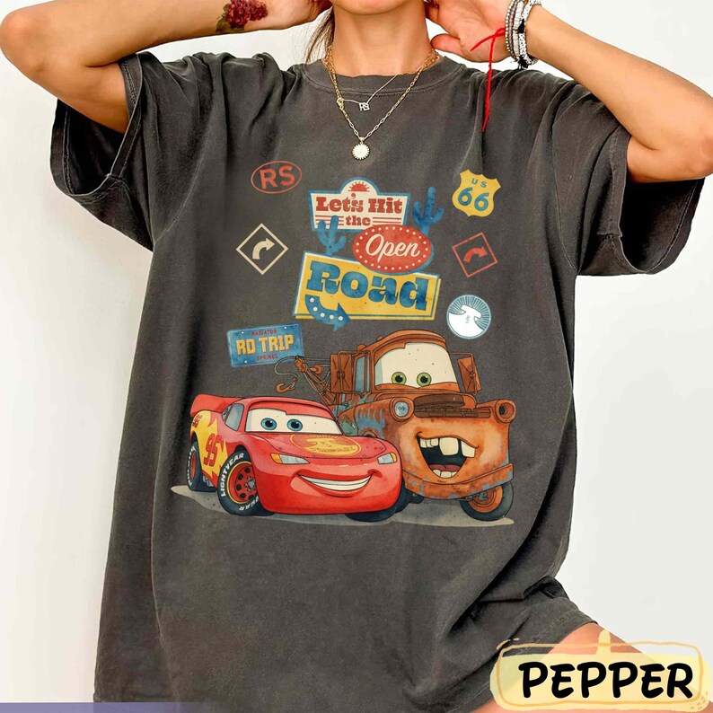 May include: Dark grey t-shirt featuring Lightning McQueen and Mater from the film Cars, with the text "Let's Hit the Open Road" and Route 66 signage. The shirt has a vintage, worn appearance.