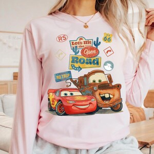 May include: A pale pink long-sleeved sweatshirt with a graphic of Lightning McQueen and Mater from the film Cars. The design includes the text "Let's Hit the Open Road" and Route 66 signage.