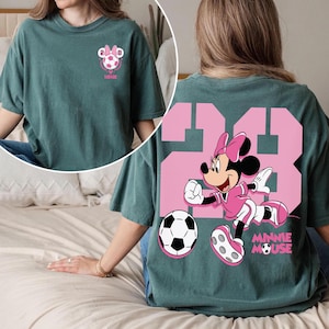 May include: Teal green t-shirt with a pink Minnie Mouse soccer design. The front has a small graphic with a soccer ball and the text "MINNIE". The back displays a large "23" with Minnie Mouse and a soccer ball, and the text "MINNIE MOUSE".