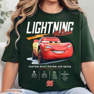 Puede incluir: Camiseta verde oscuro con Lightning McQueen, un coche de carreras rojo de la pel&iacute;cula Cars. La camiseta incluye el texto "LIGHTNING McQueen" y "95". Texto adicional detalla el motor, el chasis, la carrocer&iacute;a y el rendimiento del coche.