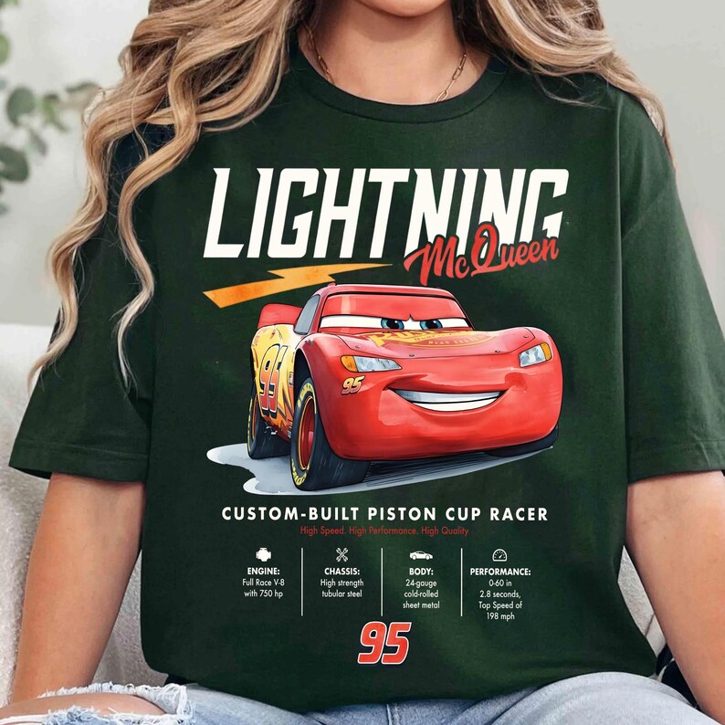 Puede incluir: Camiseta verde oscuro con Lightning McQueen, un coche de carreras rojo de la pel&iacute;cula Cars. La camiseta incluye el texto "LIGHTNING McQueen" y "95". Texto adicional detalla el motor, el chasis, la carrocer&iacute;a y el rendimiento del coche.