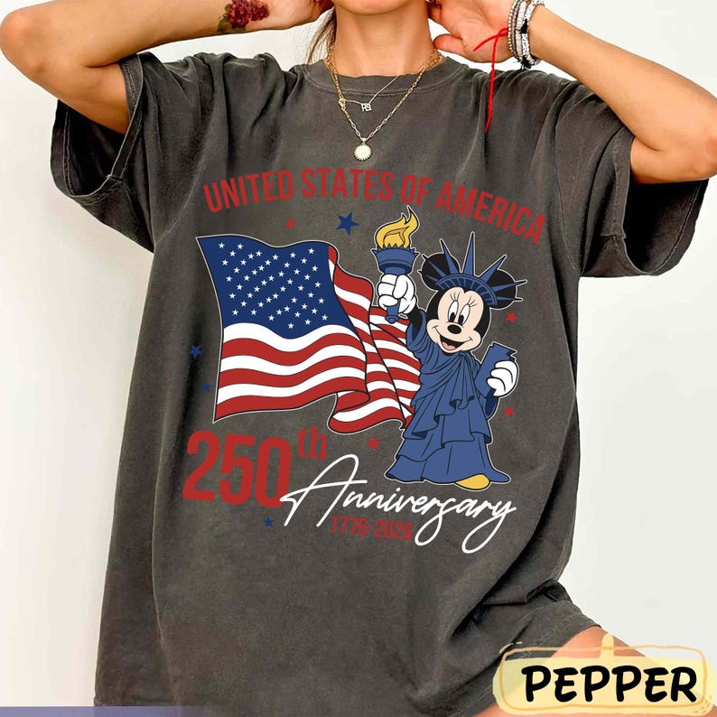 May include: Dark gray t-shirt featuring a graphic of the American flag and Minnie Mouse dressed as the Statue of Liberty. The text reads "United States of America 250th Anniversary 1776-2026".