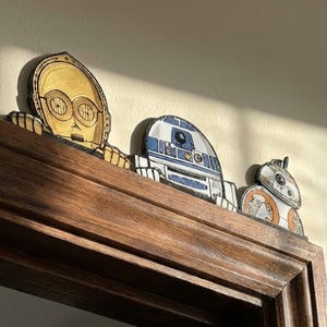 Puede incluir: Recortes de madera de los personajes de Star Wars C-3PO, R2-D2 y BB-8. Los personajes están pintados en oro, azul y blanco, y naranja y blanco. Se colocan en un marco de madera marrón oscuro, para la decoración de ciencia ficción.