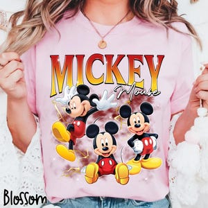 May include: Pale pink t-shirt with a graphic of Mickey Mouse in three poses. The word "Mickey" is in gold, above "Mouse" in script. The t-shirt also has the word "Blossom" in the bottom left corner.