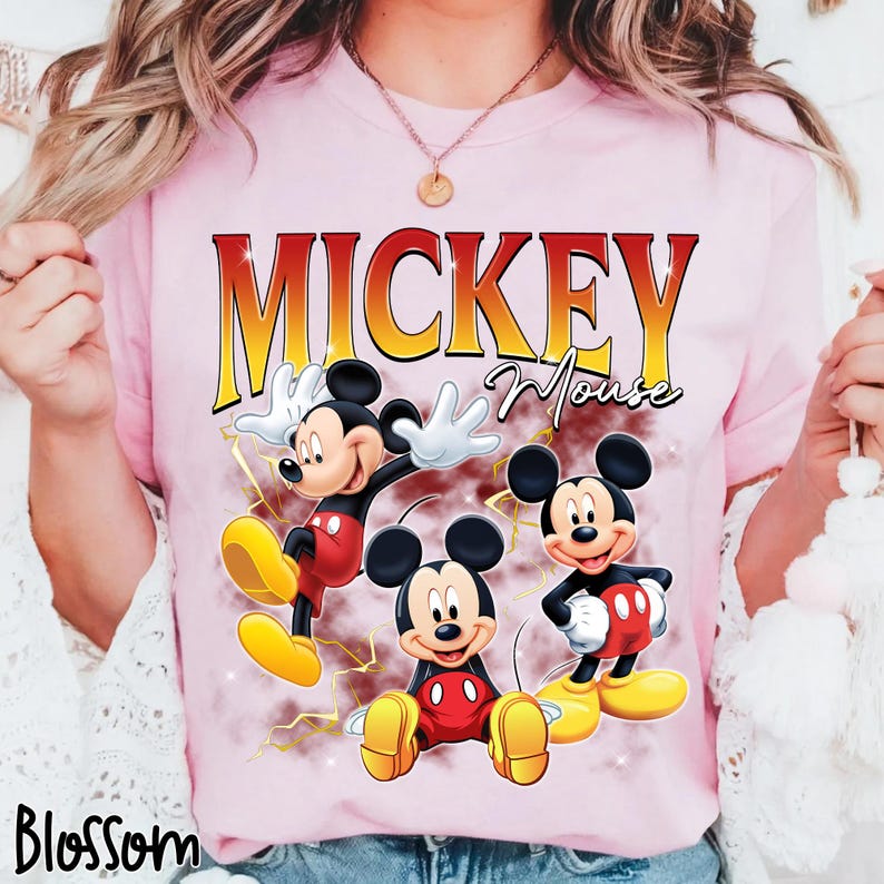 May include: Pale pink t-shirt with a graphic of Mickey Mouse in three poses. The word "Mickey" is in gold, above "Mouse" in script. The t-shirt also has the word "Blossom" in the bottom left corner.