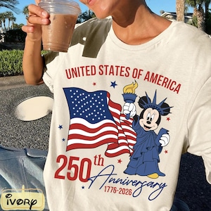 May include: Cream-colored t-shirt featuring a graphic celebrating the 250th anniversary of the United States of America. The design includes the American flag, a cartoon character dressed as the Statue of Liberty, and the text "United States of America" and "250th Anniversary 1776-2026".