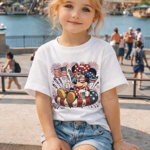 May include: A young girl wearing a white t-shirt with a graphic of Minnie Mouse in patriotic attire. Minnie is holding an American flag and a mug, surrounded by fireworks. She is wearing sunglasses with the American flag design.